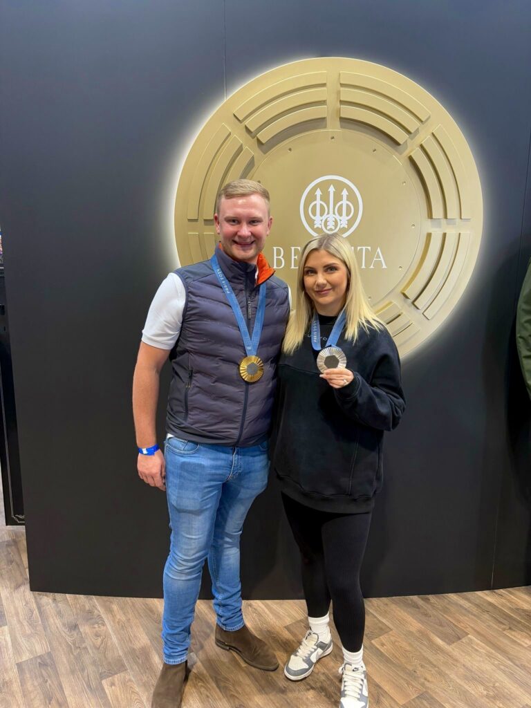 Olympic shooting medallists Nathan Hales and Amber Rutter standing at the Beretta stand, holding their gold and silver medals respectively, 