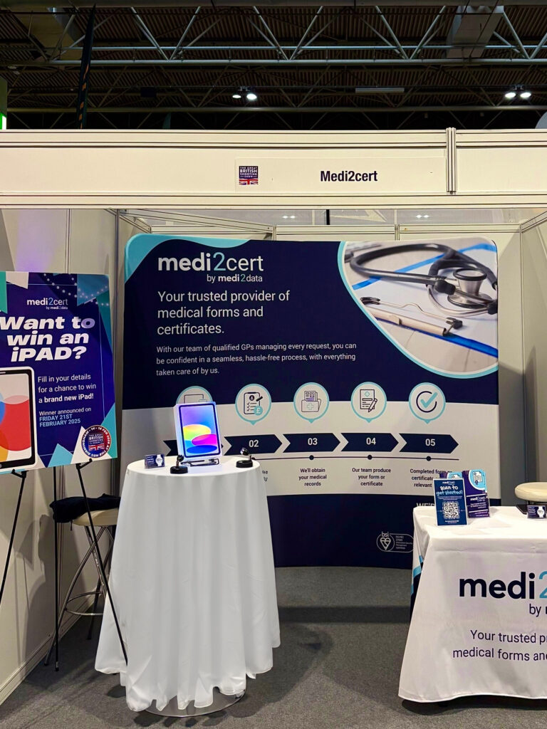 The Medi2cert stand, which has a banner in the background. In the foreground is the table with all of our flyers on, and a smaller table containing our iPad display with details about our giveaway. 